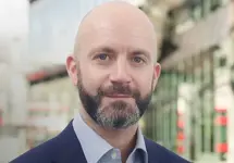 Dave Nicholson, Associate Director, Hong Kong, standing outside in front of buildings featuring an urban cityscape with a mix of architectural styles. The scene includes some windows and red features, adding a touch of color.