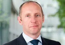 Gareth Morgan, Director, standing outside in front of an outdoor urban setting, featuring modern architectural elements with clean lines. There are hints of greenery, suggesting some trees or plants.