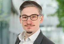 Jack Eden, Associate Director,  standing outside in front of an outdoor urban setting, featuring modern architectural elements with clean lines. There are hints of greenery, suggesting some trees or plants.