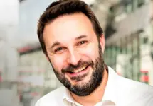 Luke De Han Project Director,  standing outside in front of buildings featuring an urban cityscape with a mix of architectural styles. The scene includes some windows and red features, adding a touch of color.