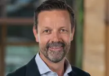 Mark McDonald, Project Director, Infrastructure, outside in front of a blurred brown background featuring a building and mix of natural and urban elements.