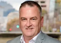 Michael Brady, Director, Qatar,  standing outside featuring an urban cityscape with tall buildings and a mix of architectural styles. The scene includes some trees with autumn foliage, adding a touch of color. 