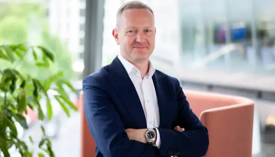 Portrait of Neil Bullen, Global Managing Director, Real Estate, In office standing by plant with his arms crossed.