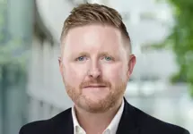 Sean Hyland, Project Director, standing outside in front of an outdoor urban setting, featuring modern architectural elements with clean lines. There are hints of greenery, suggesting some trees or plants.