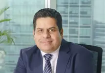 Sumit Mukherjee, Director, standing outside in front of buildings featuring an urban cityscape with a mix of architectural styles. The scene includes some windows and red features, adding a touch of color.