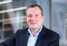 Andy Scott, Managing Director Defence, UK, standing outside in front of an outdoor urban setting, featuring modern architectural elements with clean lines. There are hints of greenery, suggesting some trees or plants.